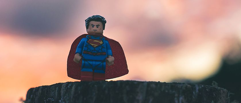 Eine kleine Lego-Figur steht auf einem Baumstumpf vor einem unscharfen, farbenfrohen Himmel. Die Figur trägt einen blauen Anzug mit rotem Umhang und einem auffälligen Brustsymbol und wirkt wie ein Superheld.