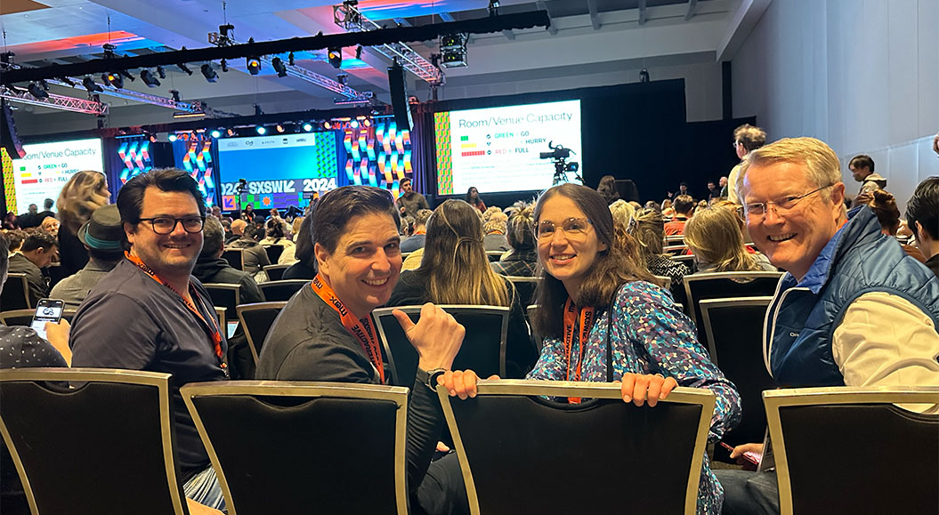 BrandTrust Gruppe von vier Personen sitzt in einem vollen Konferenzraum beim SXSW 2024 und schauen lächelnd in Richtung Kamera. Im Hintergrund ist die Bühne mit großen Bildschirmen und buntem Bühnenbild zu sehen.