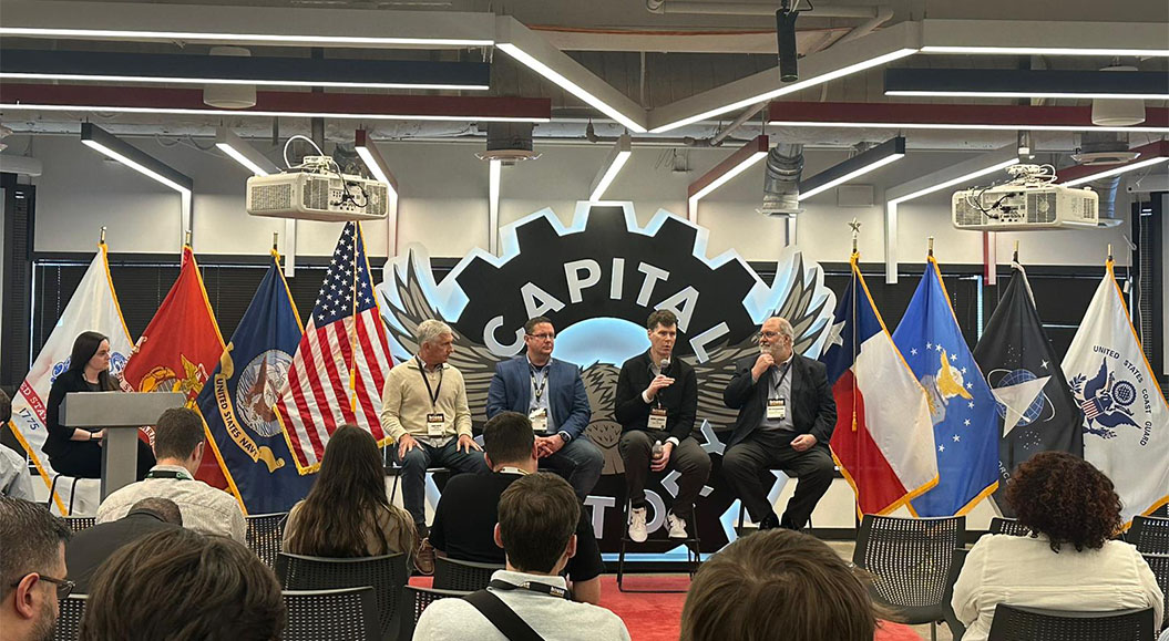 Das Bild zeigt eine Podiumsdiskussion in einem modernen Veranstaltungsraum. Auf der Bühne sitzen fünf Personen, darunter Vertreter aus verschiedenen Bereichen, die an einem Panel teilnehmen. Zu sehen sind mehrere Flaggen, darunter die der USA, Frankreichs und des US-Militärs. Eine Frau ist am Pult zu sehen, die vermutlich die Diskussion moderiert oder Fragen stellt. Im Hintergrund ist ein großes Logo zu erkennen, das "CAPITAL" und ein stilisiertes Flügel-Design zeigt. Das Publikum sitzt in Stuhlreihen und blickt auf die Bühne.