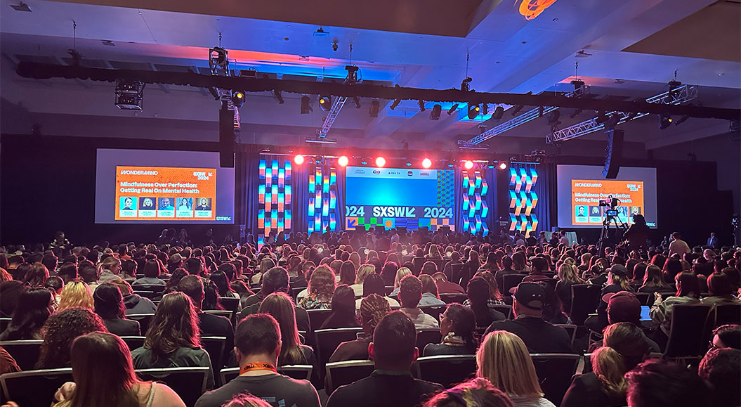 Blick auf eine große Konferenzbühne mit einer riesigen Menschenmenge im Publikum. Die Bühne ist lebendig beleuchtet mit roten und blauen Lichtern. Auf dem Bildschirm wird das Thema 'Mindfulness Over Perfection: Getting Real On Mental Health' angezeigt, zusammen mit den Namen der Teilnehmer. Es handelt sich um eine Veranstaltung im Rahmen des SXSW Festivals 2024.