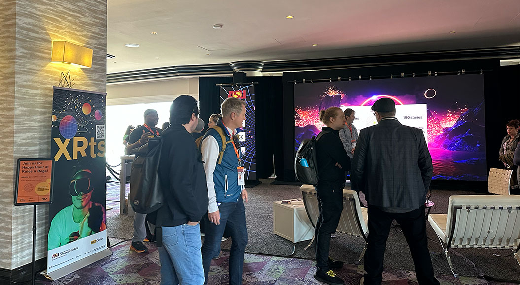 eine Gruppe von Personen, die an einer Veranstaltung oder Messe teilnehmen. Es gibt eine Bannerwerbung im Vordergrund, die das Wort „XRTs“ zeigt, zusammen mit einem Bild einer Person, die eine VR-Brille trägt. Im Hintergrund ist eine große Projektionswand zu sehen, die eine futuristische Landschaft mit leuchtenden Farben darstellt. Weitere Personen stehen oder gehen in der Nähe und es scheint, als ob eine Aktivität oder Präsentation stattfindet. Der Raum ist modern und gut beleuchtet. Ein zusätzliches Schild wirbt für eine Happy Hour.