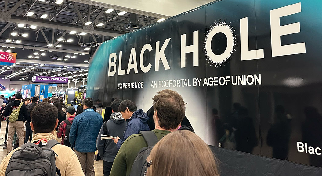 Menschen stehen in einer Schlange vor einer Wand mit dem Schriftzug „BLACK HOLE EXPERIENCE“ und dem Hinweis „AN ECOPORTAL BY AGE OF UNION“. Im Hintergrund ist ein Bereich mit verschiedenen Ständen zu sehen, darunter der „Korea Pavilion“. Einige Besucher tragen Rucksäcke und Jacken, während sie auf die Teilnahme an der Erfahrung warten.