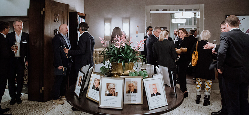 In einem eleganten Raum mit einer Gruppe von Menschen, die sich unterhalten und sich auf einem Event befinden. Einige Personen stehen an einer Wand, andere in der Mitte des Raumes, während sie Notizbücher halten und sich mit Getränken in der Hand unterhalten. Auf einem Tisch in der Mitte sind mehrere Porträtbilder in Bilderrahmen ausgestellt. Die Blumen im Vordergrund und die dezenten Lichtelemente verleihen dem Raum eine warme, gehobene Atmosphäre.