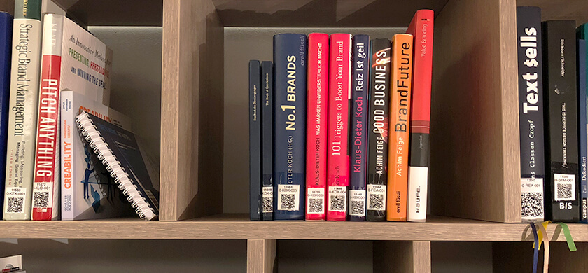 BrandTrust Bibliothek; Ein Regal mit verschiedenen Fachbüchern und einem Notizbuch. Die Bücher sind thematisch auf Markenführung, Business und Kreativität ausgerichtet. Zu sehen sind Titel wie „No. 1 Brands“, „101 Triggers to Boost Your Brand“, „Good Business“, „BrandFuture“ und „Text Sells“. Das Notizbuch mit einem Spiralring ist auf der linken Seite des Regals zu sehen, und alle Bücher sind mit QR-Codes versehen.