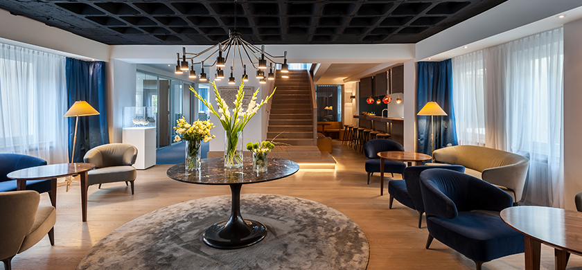 BrandTrust Büro in Nürnberg, die German Design award gewonnen hat: Ein elegantes, modernes Lounge-Ambiente mit einem runden Tisch im Vordergrund, umgeben von bequemen Sesseln und Sofas in Blau und Beige. Auf dem Tisch stehen mehrere Glasvasen mit frischen Blumen. Die Wände sind mit langen, weißen und blauen Vorhängen dekoriert. Im Hintergrund ist eine Treppe zu sehen, die zu einem weiteren Bereich führt, sowie eine Bar mit hohen Stühlen und leuchtenden Pendelleuchten. Das Raumdesign ist minimalistisch, mit Holzboden und warmem Licht.