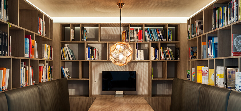BrandTrust Bibliothek: Ein moderner, gut beleuchteter Arbeitsbereich in einer Bibliothek mit Regalen voller Bücher. In der Mitte des Raumes befindet sich ein Tisch mit zwei gepolsterten Sitzen. Über dem Tisch hängt eine geometrische Pendelleuchte. Auf dem Tisch steht ein Monitor. Die Regale sind ordentlich angeordnet und enthalten eine Vielzahl von farbigen Buchrücken, die auf Fachliteratur hinweisen.