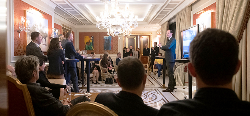 Eine elegante Veranstaltung in einem luxuriösen Raum mit Kronleuchtern und stilvollem Interieur. Im Vordergrund sind mehrere Personen zu sehen, die in einer lockeren Haltung stehen und dem Vortrag eines Mannes auf der Bühne zuhören. Der Vortragende und Brandtrust Gründer Klaus-Dieter Koch gestikuliert und spricht vor einer Präsentation, während die Gäste in kleinen Gruppen oder an Stehtischen verteilt sind. Einige Personen sitzen auf bequemen Stühlen und beobachten das Geschehen.