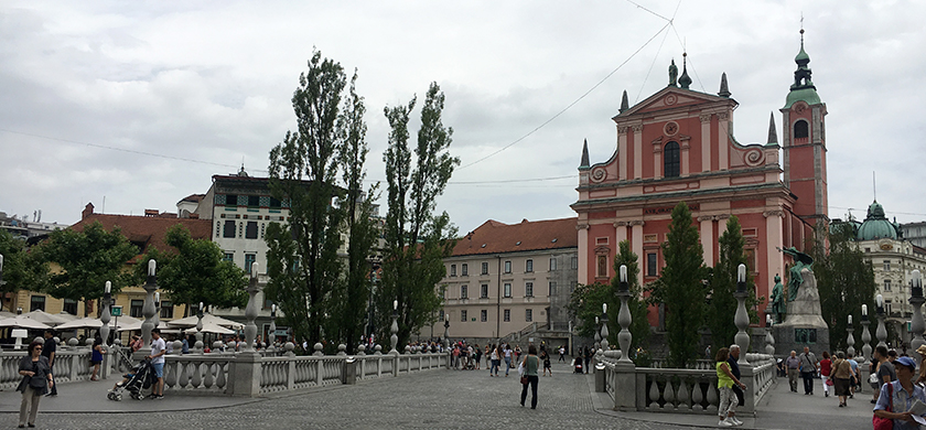 BrandTrust in Slowenien: Blick auf den Prešeren-Platz in Ljubljana mit der pinken Franziskanerkirche im Hintergrund. Der Platz ist von einer Fußgängerbrücke umgeben, auf der mehrere Menschen unterwegs sind. Bäume und Cafés säumen den Rand des Platzes, während im Vordergrund ein Denkmal zu sehen ist.